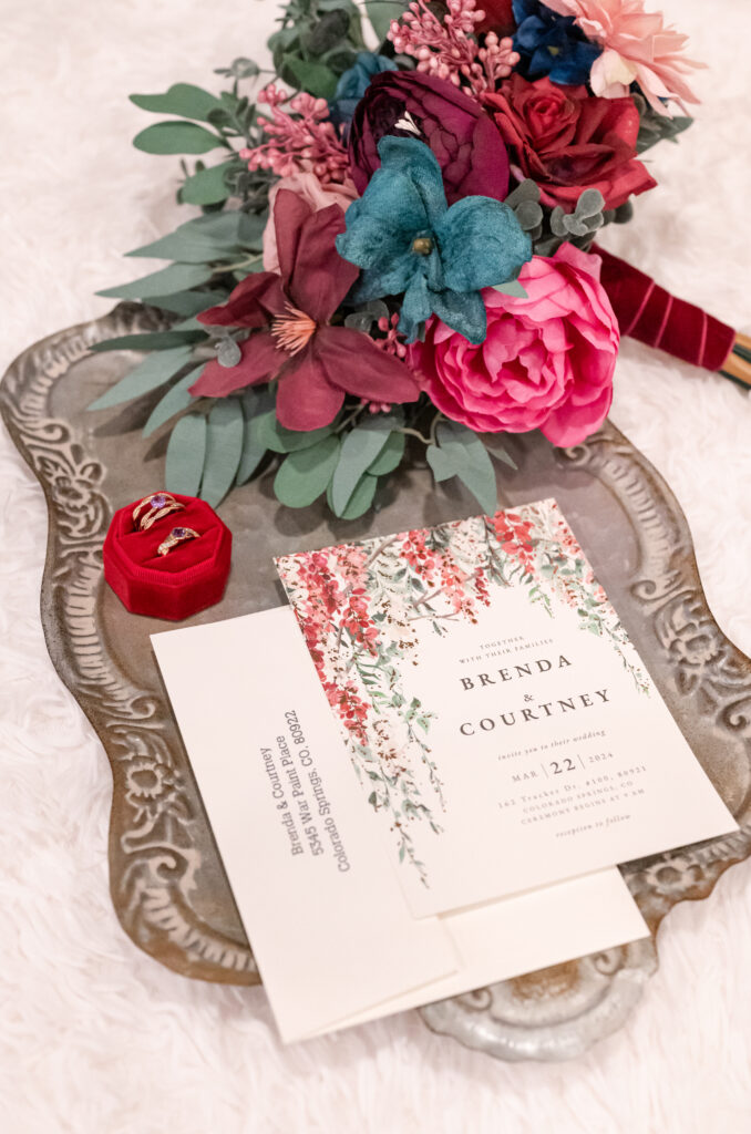 Wedding invitations laying on a gray vintage tray with flower bouquet and wedding rings