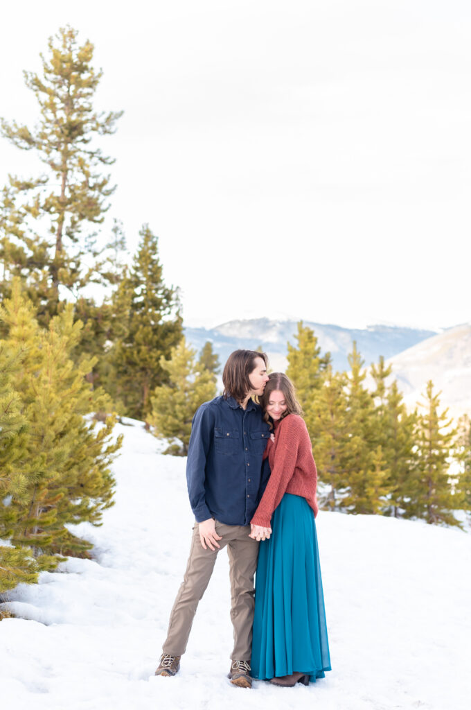 Young man kisses his bride-to-be as they stand in the snow against a background of evergreen trees and distant mountains while they pose for engagement photos in Colorado