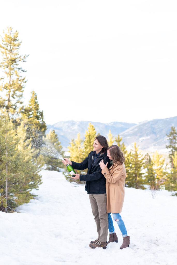 Man and woman open a bottle of champagne to celebrate their engagement in the snowy mountains of Colorado