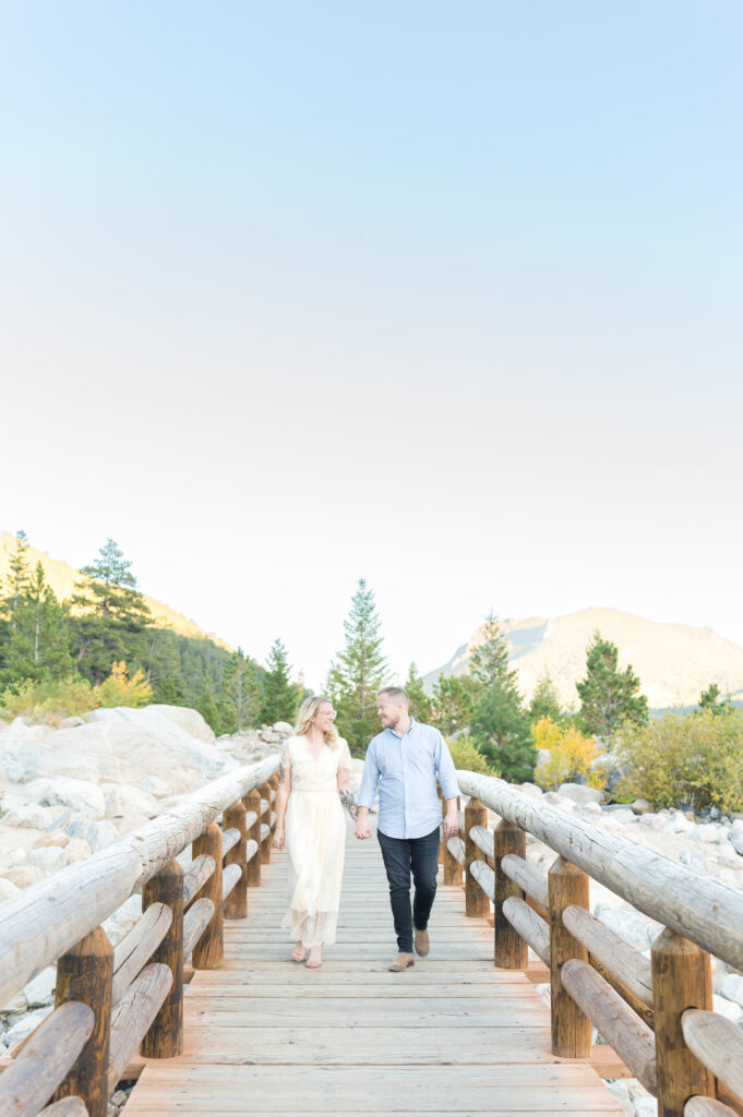 Woman in white dress and man in button-up shirt and slacks crossing a rustic wood bridge in Colorado Springs