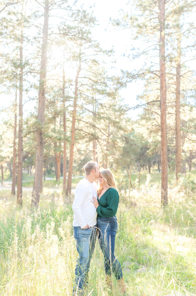 Young man kisses his sweetheart's forehead as they stand together in a wooded park