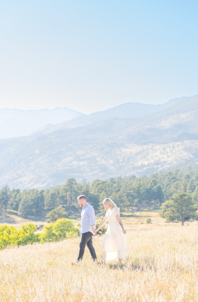 Couple holding hands as they walk through a sunlit field with striking mountain views in the background
