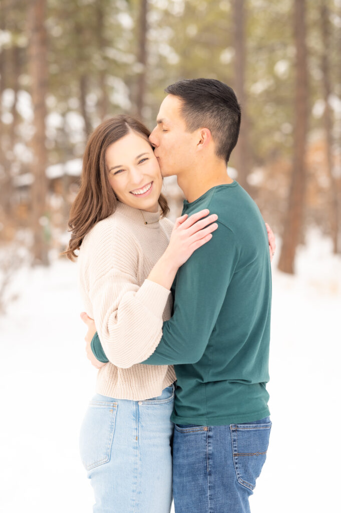 Young man kisses his fiancee's cheek as she puts her arms around him and smiles