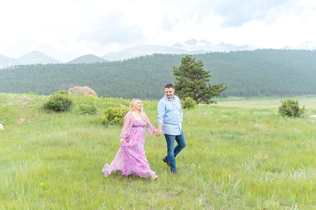 Woman in long floral dress strides through a Colorado meadow holding her fiance's hand during their engagement photoshoot