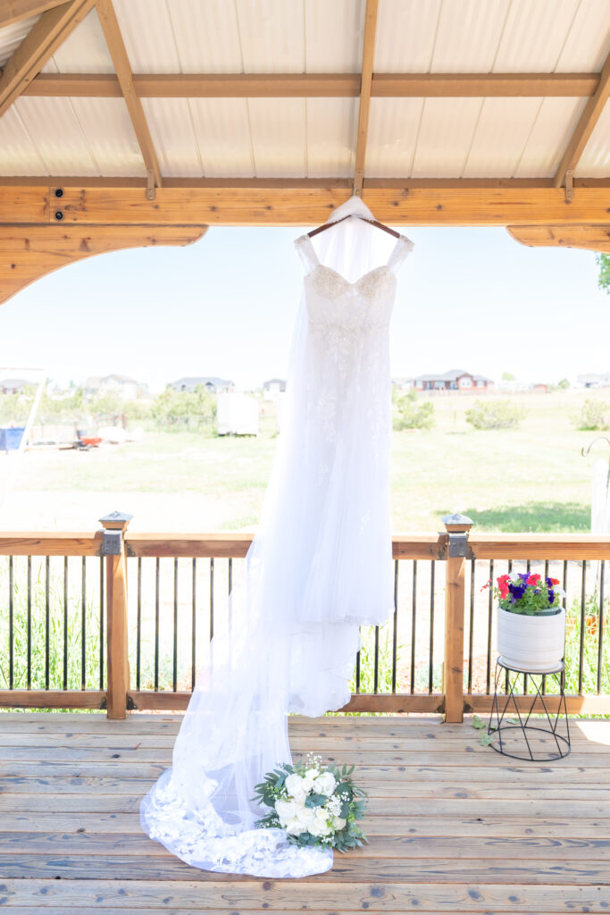 Detail photo of the bride's dress hanging up and the wind beautifully blowing the veil with the bride's flower bouquet laying on her dress