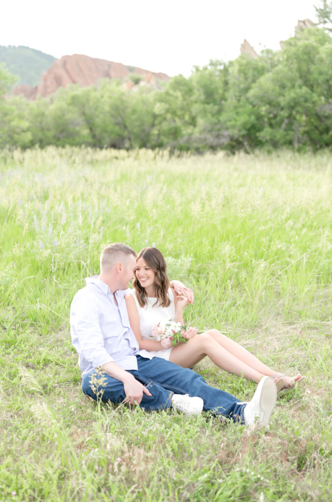 Couple sitting together in the grassy field while he is putting his arm around her and she is holding flowers in her lap