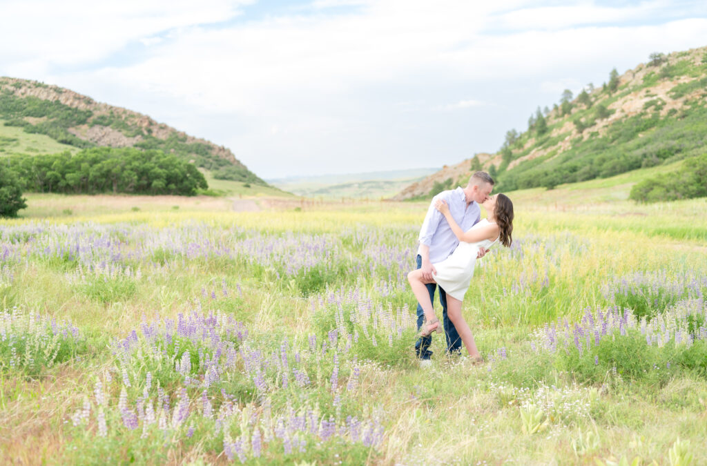 Couple sharing a romantic dip kiss in mountain meadow grassy field with purple wildflowers at Roxborough State Park in Colorado