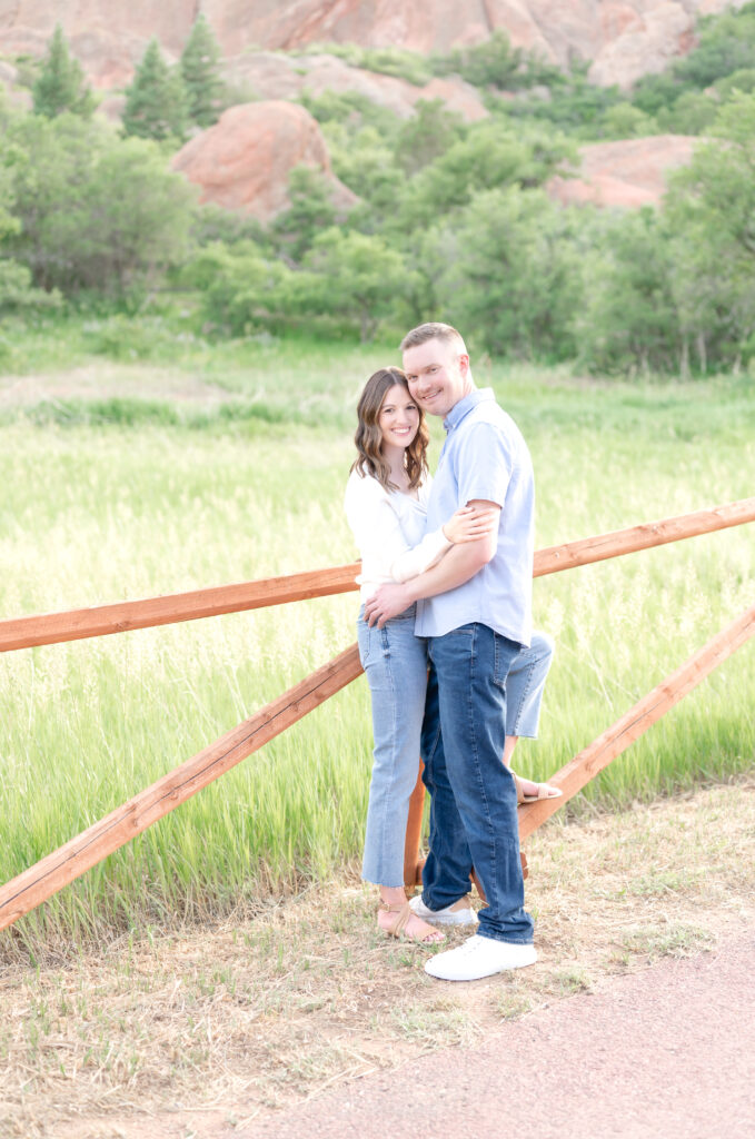 Couple leaning into a brown fence at Roxborough State Park while smiling towards the camera