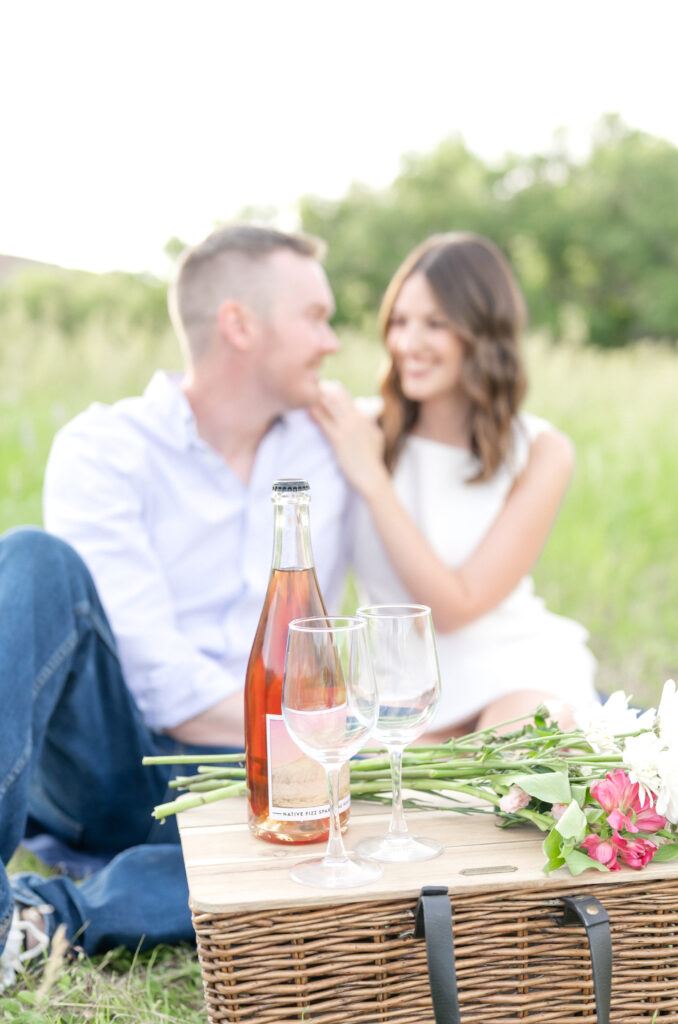 Girl putting resting her hands on his shoulder while looking and smiling at each other with picnic basket and wine sitting on ground near them