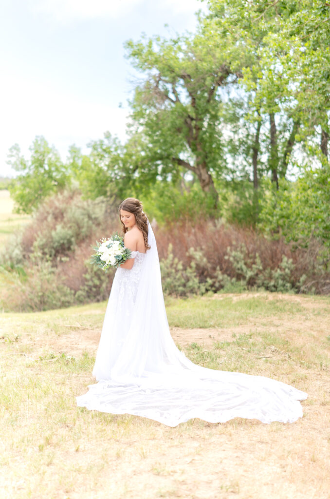 Bride standing in field looking down over her shoulder while holding her bridal bouquet and the train of her dress laid out