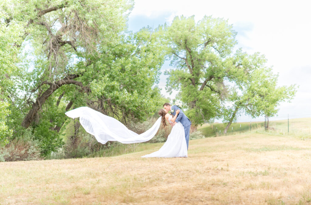 Wide photo of the groom dipping his bride back for a romantic kiss on their wedding day with the bride's veil flowing in the wind