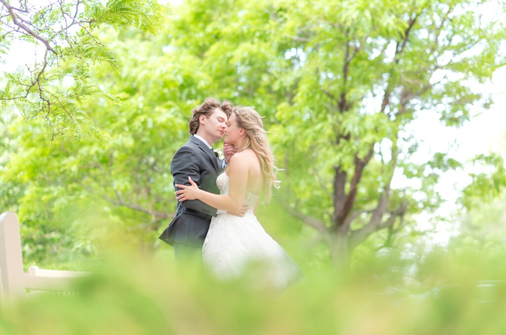 Bride and groom leaning in for a kiss with greenery foreground and background