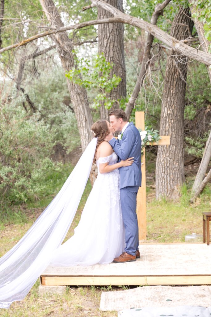 Bride and groom sharing their first kiss as husband and wife at their Colorado wedding ceremony site with wooden rustic cross behind them 