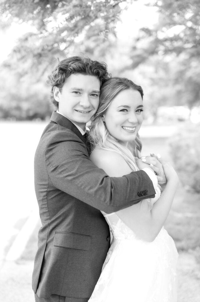 Groom hugging his wife from behind and both smiling towards the camera