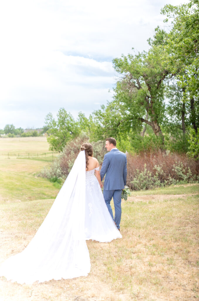 Bride and groom walking away out into an open field holding hands together with the bride's veil laid out behind her 