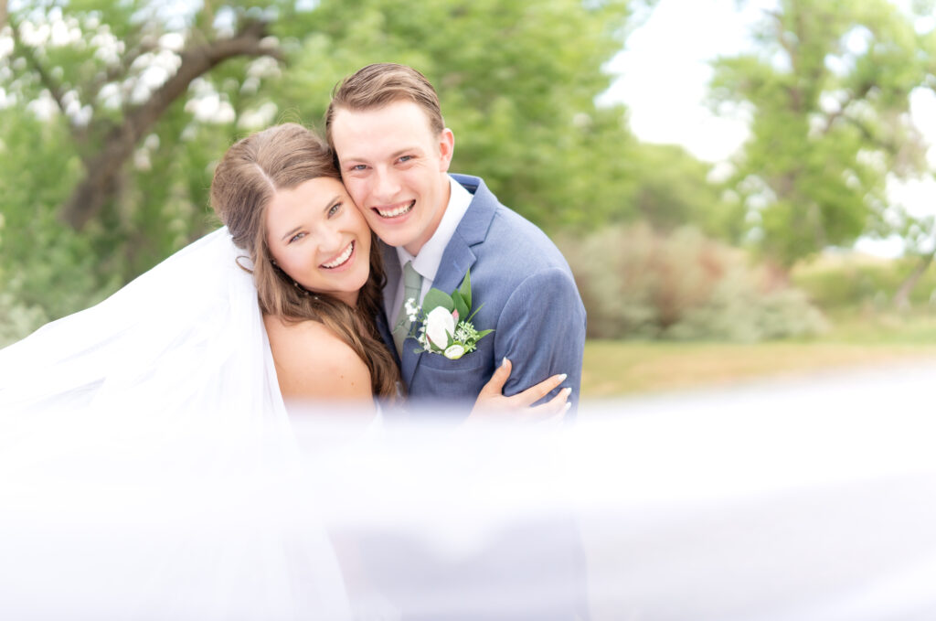 Bride and Groom leaning in towards camera together laughing and smiling with bride's veil flowing in the wind