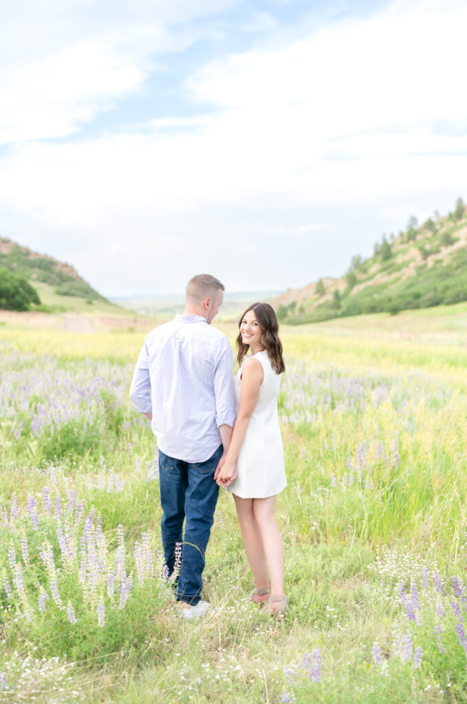 Couple walking and holding hands in grassy wildflower field while the girl is looking back and smiling towards the camera