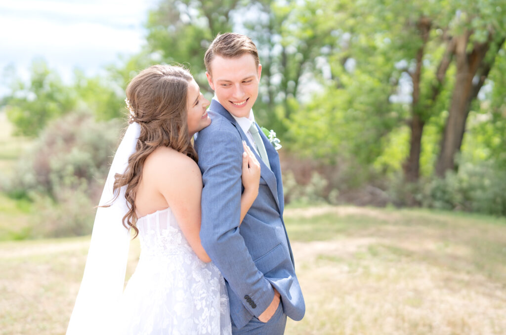 Bride hugging groom from behind as they stand in a clearing together