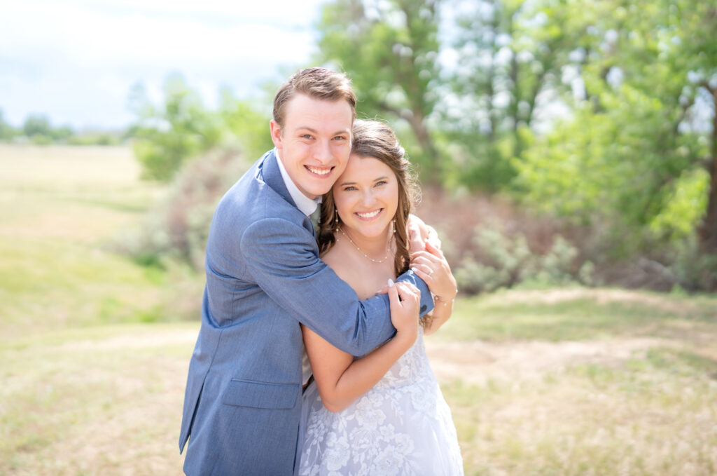 Groom wraps his arms around his bride from behind as they both smile at the camera