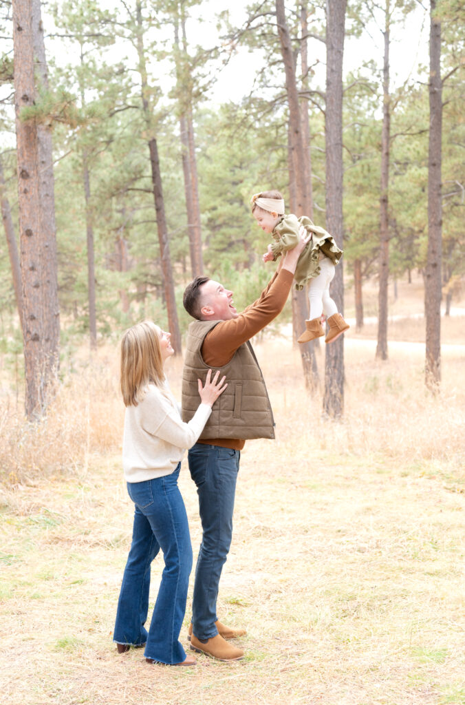 Dad lifts his baby daughter up into the air, laughing, while mom looks on and smiles