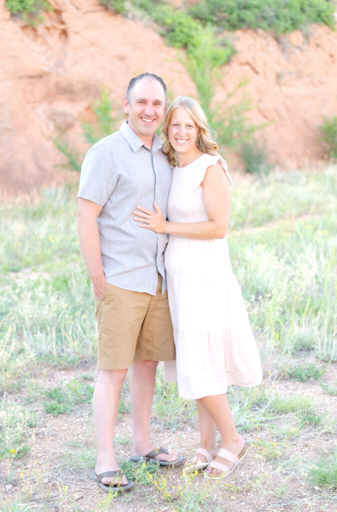 Man in light-colored short-sleeve button-up shirt and khaki shorts puts his arm around his wife wearing a white sundress