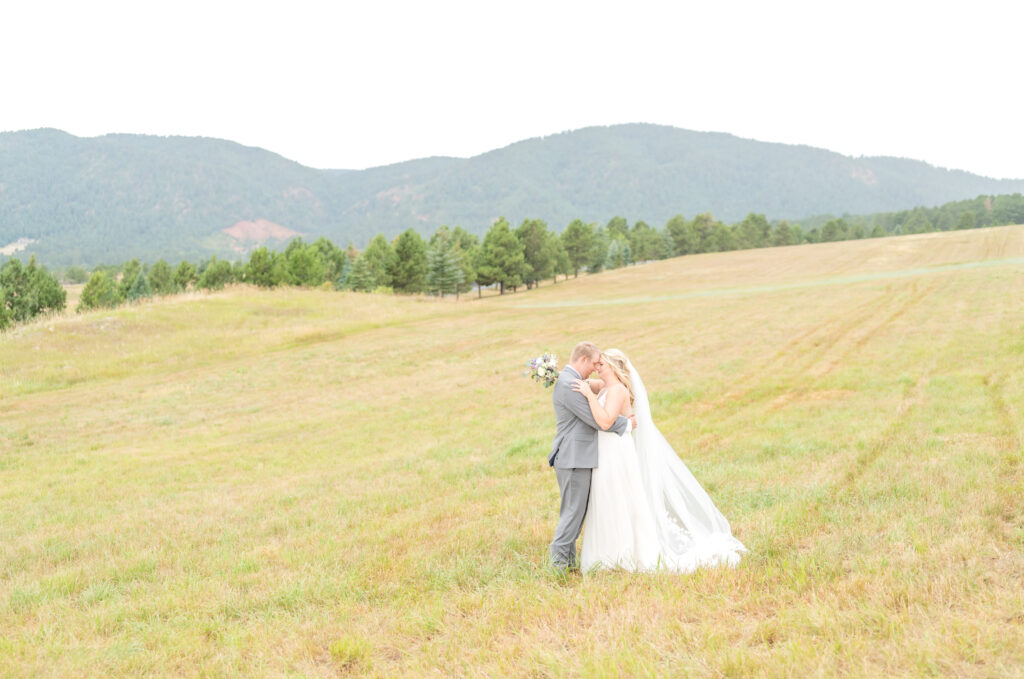 Couple in their wedding clothes hold each other close as they pose in a field with a beautiful mountain backdrop