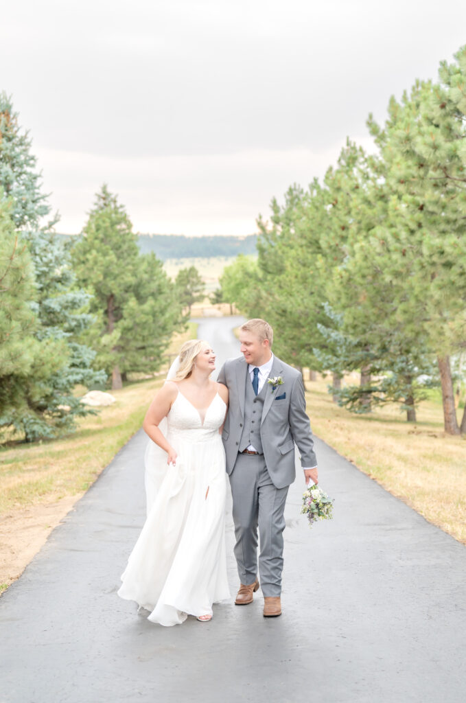 Newly married couple laughing as they walk together up the tree-lined path of a mountain ranch