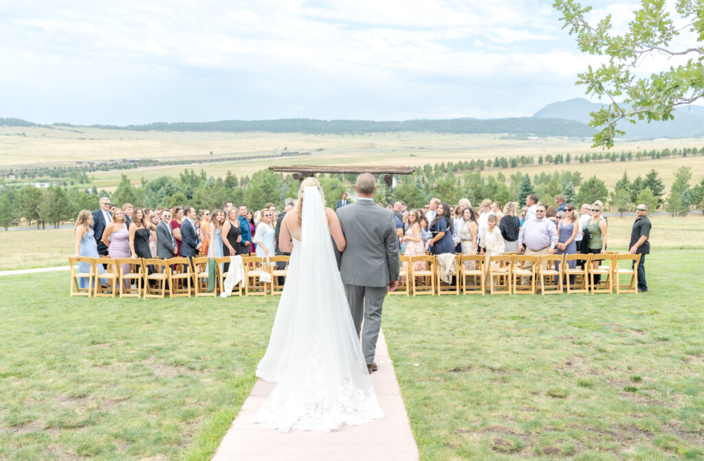 Bride holds her father's arm as they walk down the path together towards the wedding guests seated in an outdoor venue