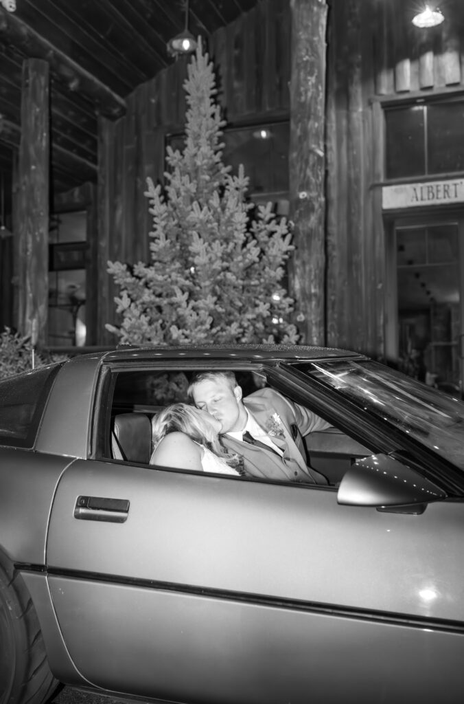 Black and white flash photo of bride and groom seated in retro car exchanging a kiss