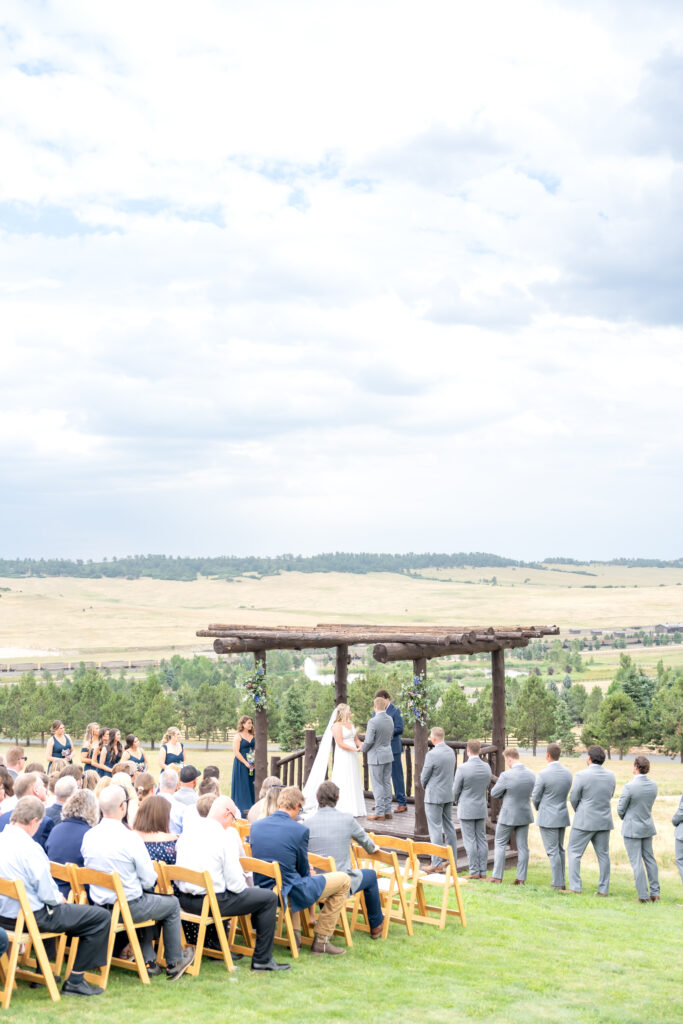 Bride and groom holding hands in a rustic log arbor while their guests look on