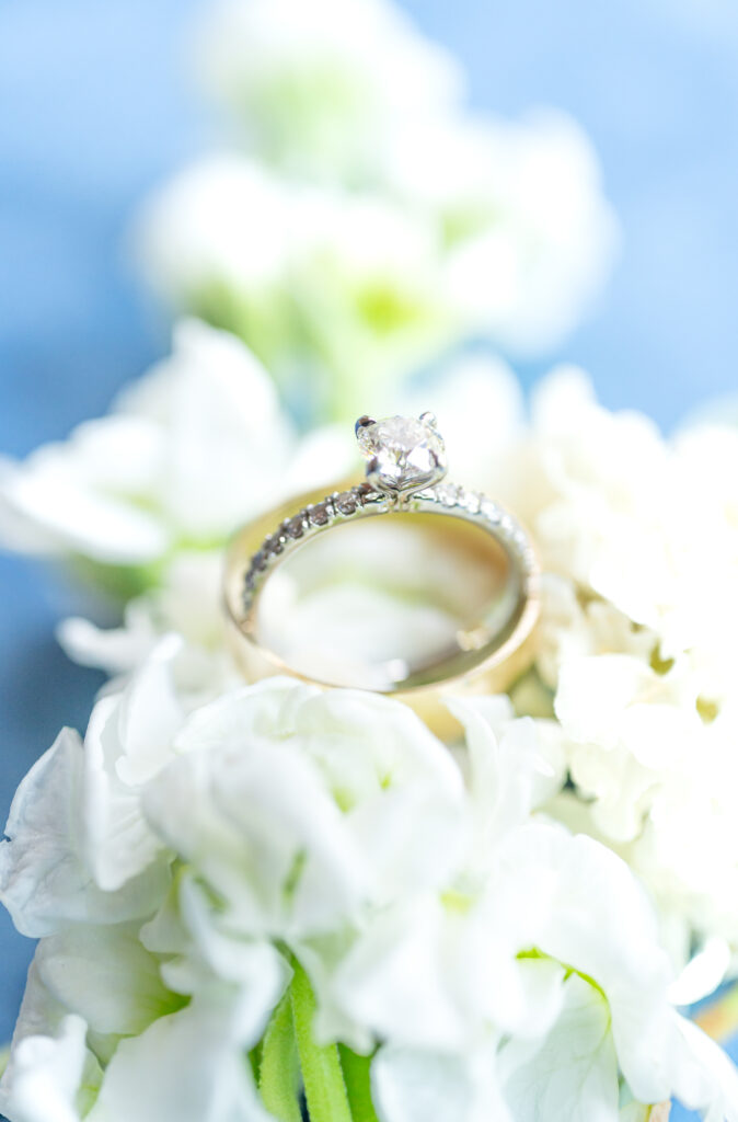 Closeup of bride's wedding ring arranged on a bed of white flowers against a blue backdrop