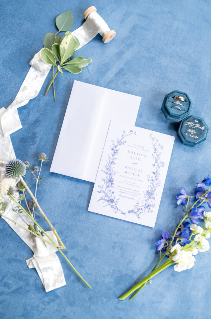 Wedding invitations and rings on a blue cloth surrounded by flowers and a spool of ribbon