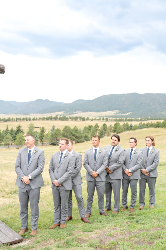 Groomsmen in gray suits and blue ties watch the wedding ceremony against a backdrop of mountains and blue sky