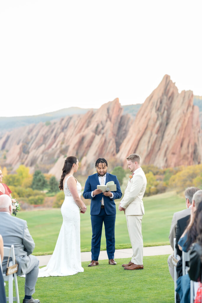 Bridge and groom stand on either side of their officiant during their wedding at Arrowhead golf course with red rock formations towering behind them