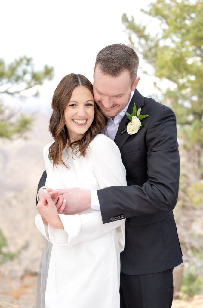 Groom holding his wife in a tight snuggle hug while looking down over her shoulder at her smiling 