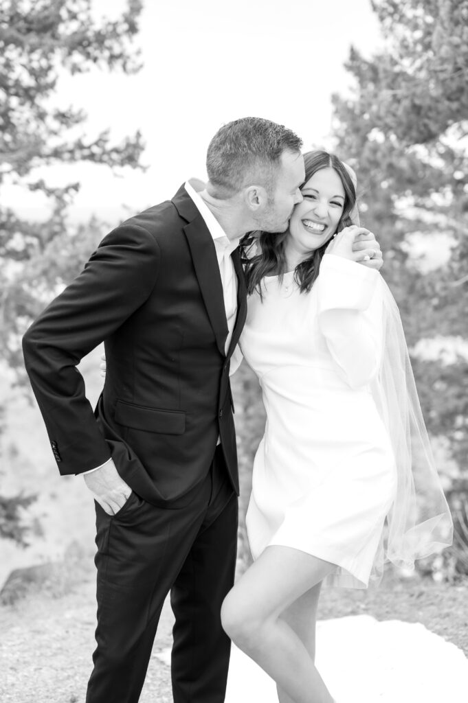 Groom pulling his bride in close for a kiss on the cheek while smiling and laughing together during their outdoor elopement ceremony at Lookout Mountain in Colorado