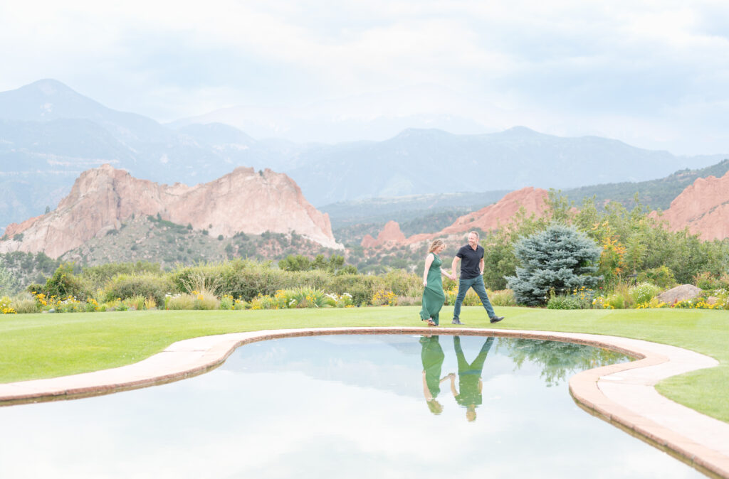 Couple hold hands as they stroll on a manicured lawn near a pond with mountain views in the background
