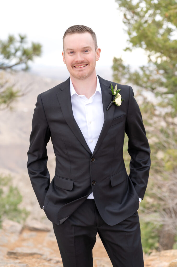 Groom smiling towards the camera with his hands in his pockets with backdrop of red rock landscape and trees behind him