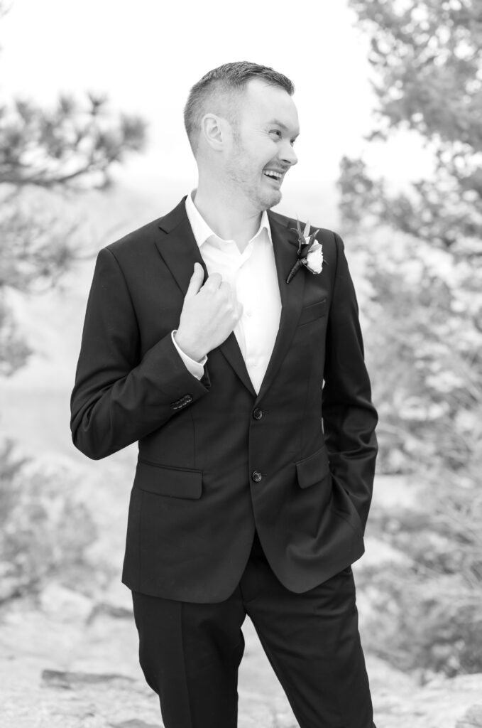 Groom holding the edge of his black suit jacket while looking over to the side smiling and laughing on his elopement day in Colorado