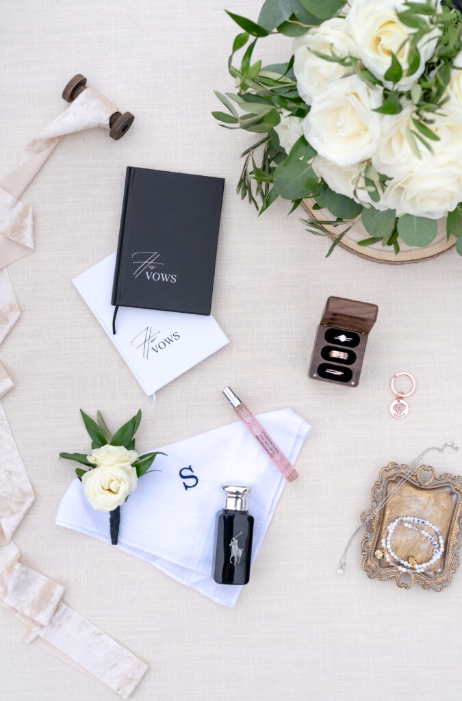 Wedding vows, rings, florals, ribbon, perfume, and cologne all beautifully laid out and arranged on a cream backdrop mat