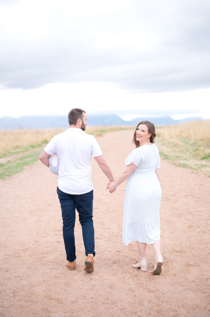 Young couple hold hands as they walk along a sandy path in Colorado Springs while Dad holds baby in one arm and Mom looks back over her shoulder and smiles