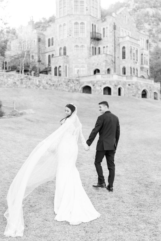 Bride and groom hold hands as they walk up the manicured lawn towards Glen Eyrie Castle in Colorado Springs
