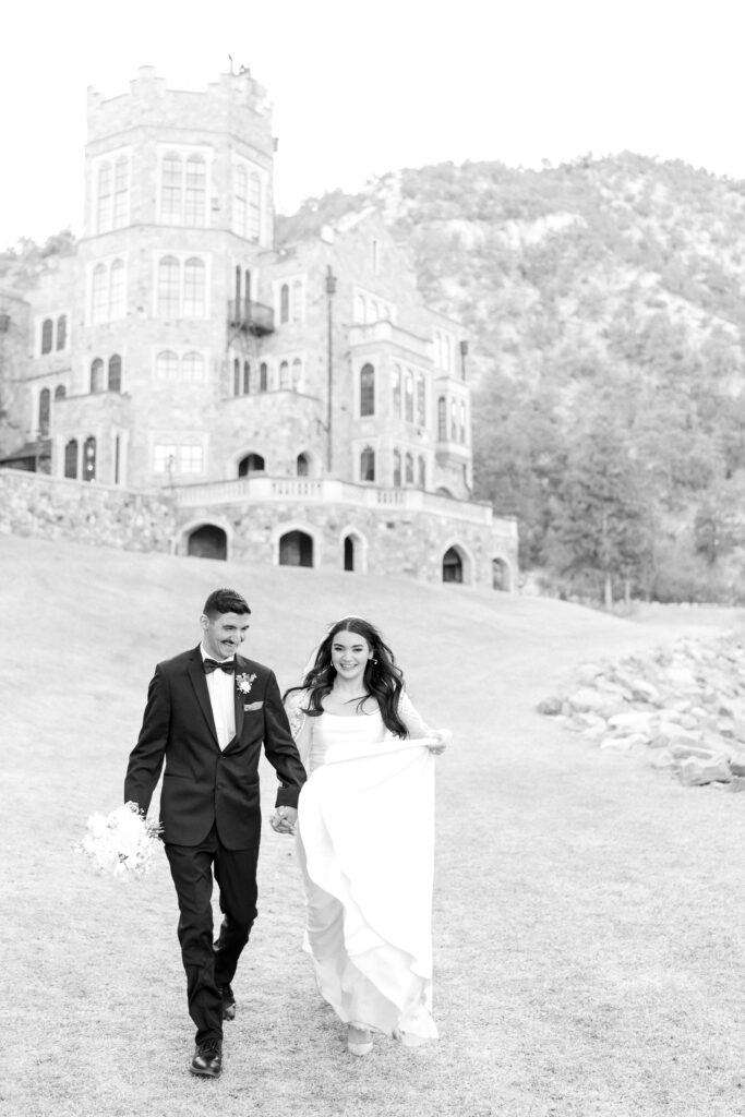 Bride and Groom running and laughing together while holding hands with glen eyrie castle behind them