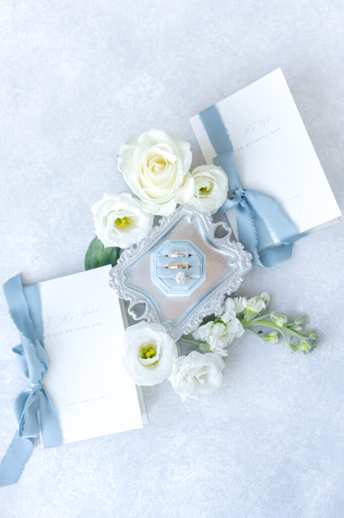 Wedding detail photo of wedding vow books, white florals and greenery, and blue velvet ring box of the rings