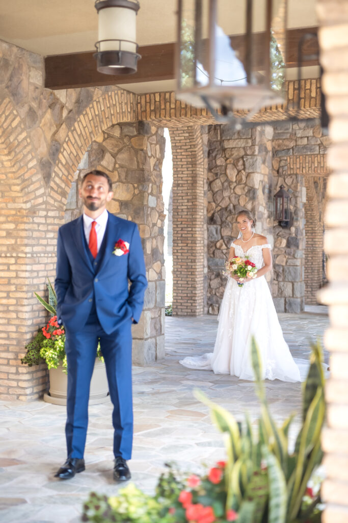 Groom standing in stone entryway with his hands in his pockets while his bride walks towards him from behind during their wedding first look