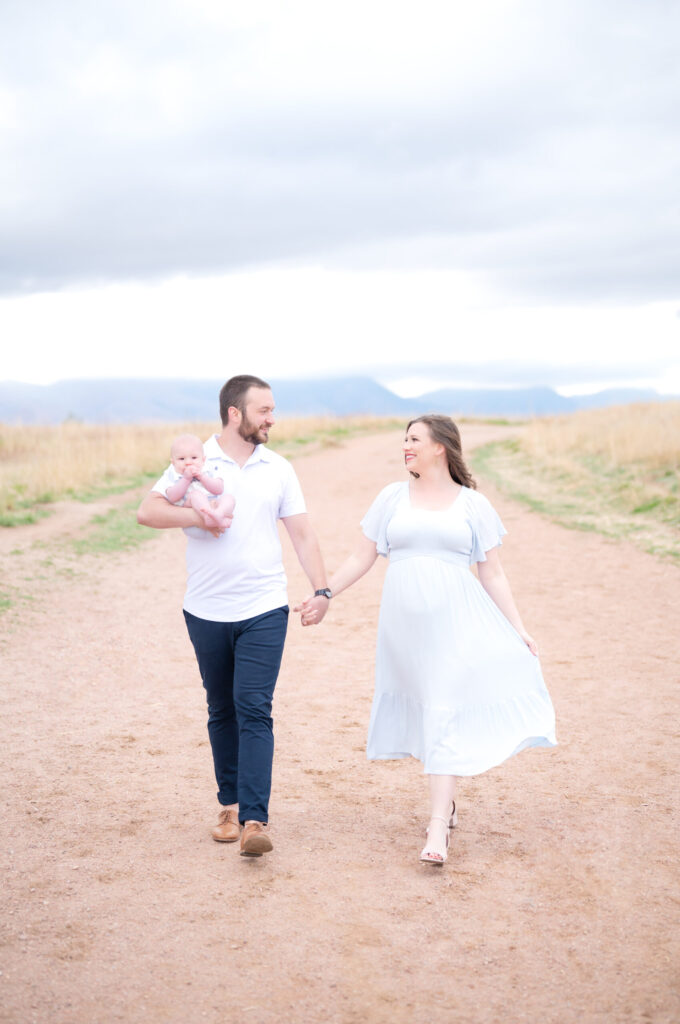 Young couple walk hand in hand down a dirt road in Colorado Springs, dad carrying baby in the crook of his arm