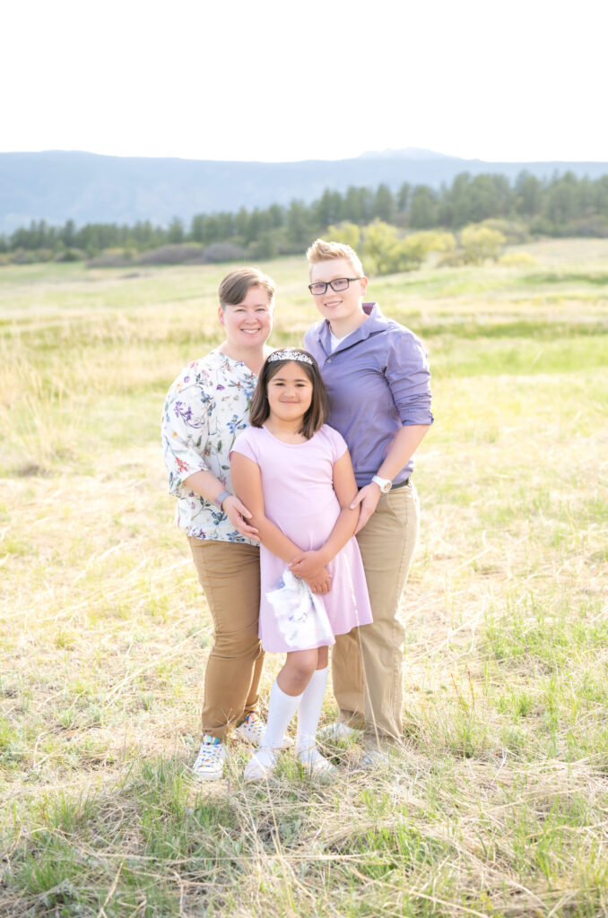 Two moms smiling with their daughter in open grassy field during their Colorado Springs family photoshoot