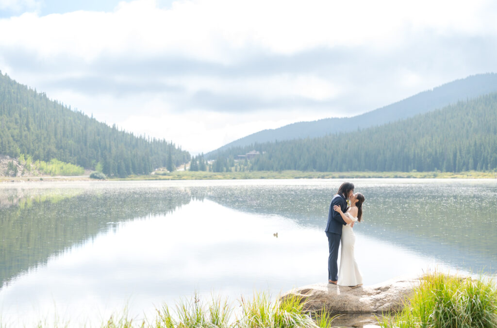 Bride and groom exchange a kiss as they stand together on a rock in Echo Lake with the sun shining down at them and the mountains reflected in the water behind them
