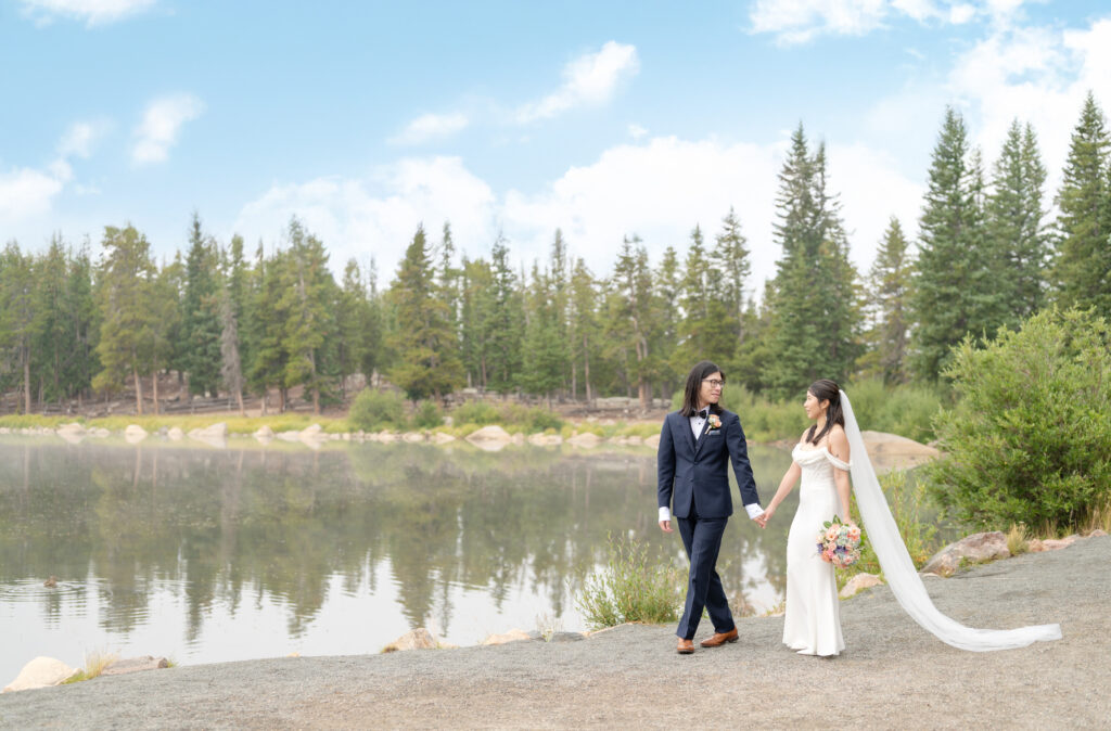 Couple in wedding clothes strolling hand in hand along the edge of a calm lake on an overcast morning in Colorado