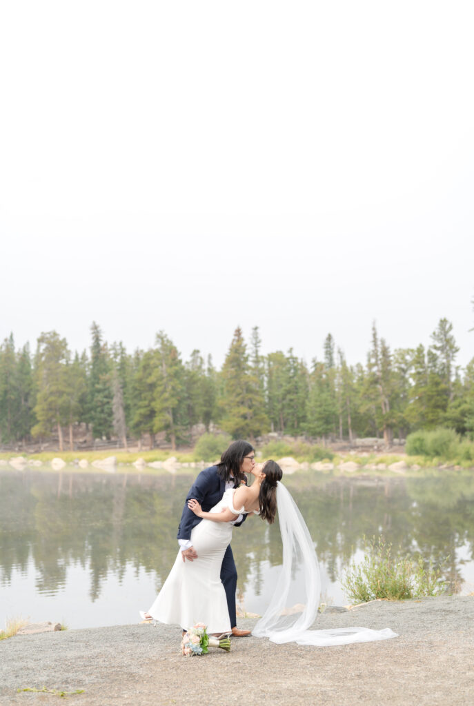 Groom dips his bride for a kiss as they stand together on the edge of Echo Lake with evergreen trees mirrored in the water behind them